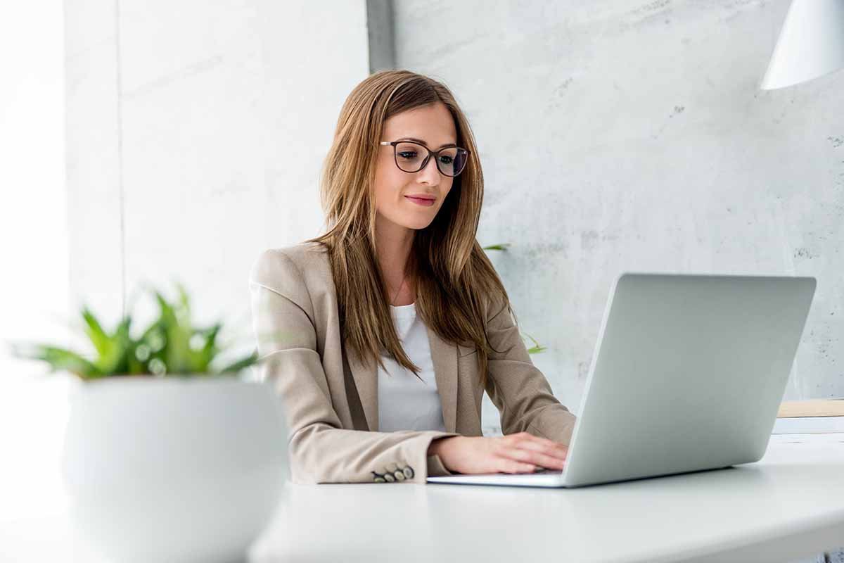 A young lady wearing glasses working on her laptop dressed in a tan suit jacket and white blouse.
