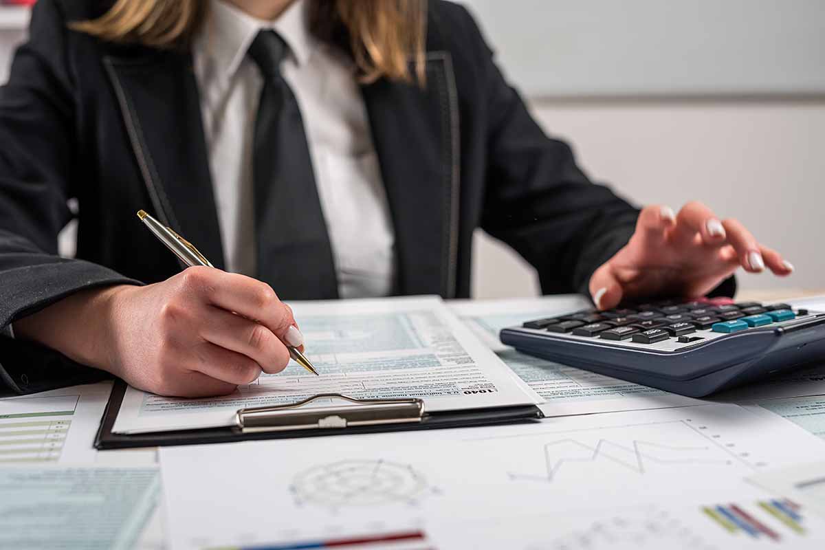 A womna in a business suit using a calculator and writing on a clipboard.