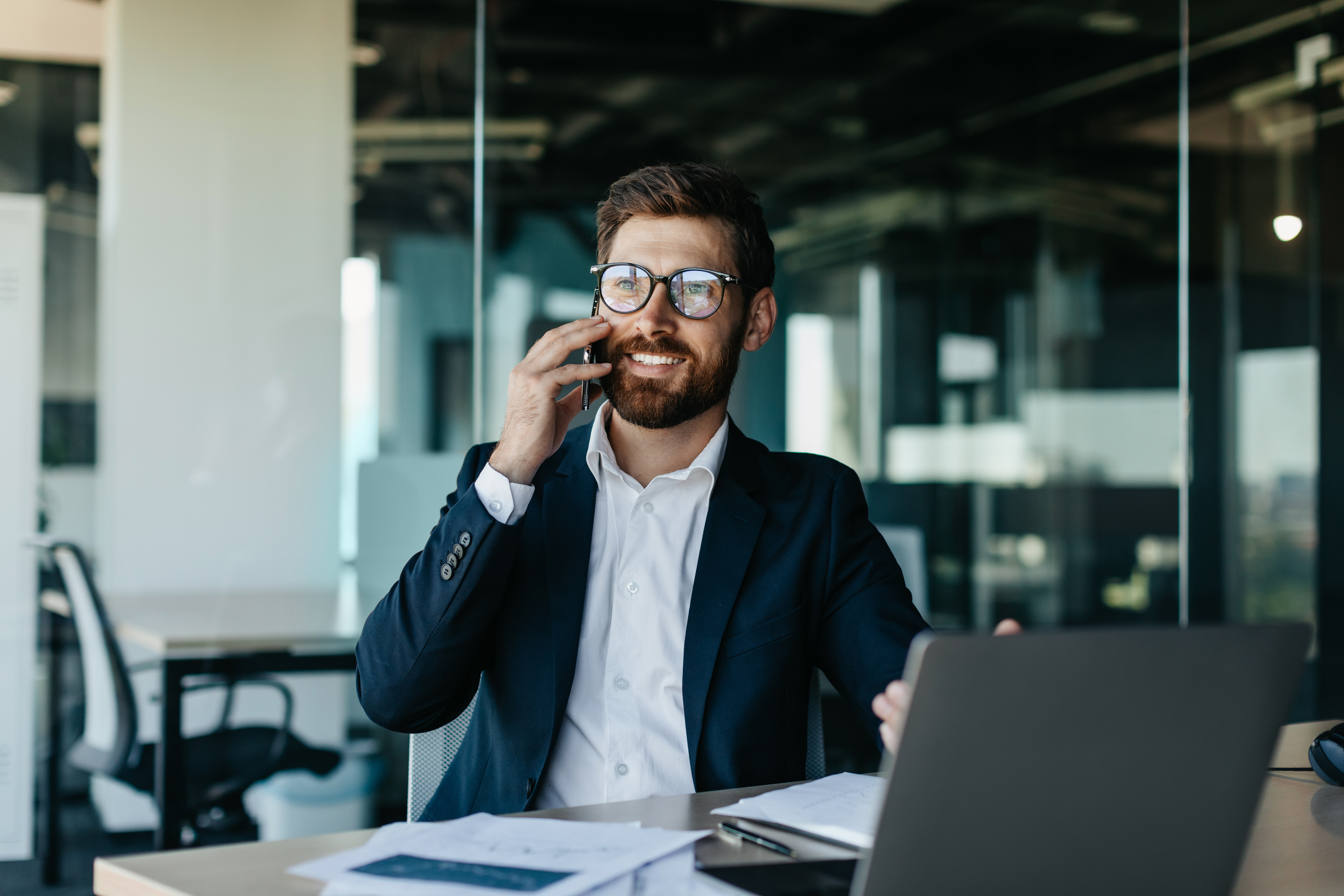 Business professional in a modern office smiling while talking on the phone, with documents and a laptop on the desk – representing communication, success, and corporate operations.